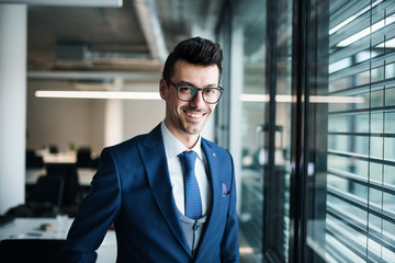 A portrait of young businessman standing in an office, looking at camera.