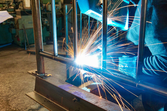 A welder at work in a workshop produces metal structures. Sparks from welding fly around.
