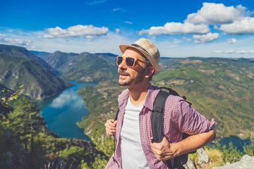 Naklejka premium Fashionable backpacker on cliff against lake or river surrounded by hills.