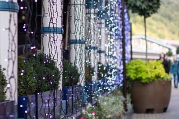 Beautiful view from the street to the cafe in the greenery of trees and flowers. Street cafe decorated with lights for the holiday.
