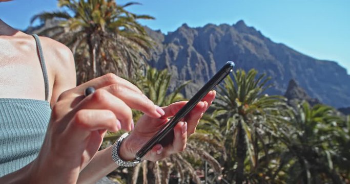 Close-up young woman swiping on smartphone social news outdoor. Tourist with cellphone on viewpoint in Masca valley tropical landscape on wonderful day. Tenerife, Spain.