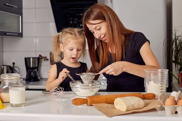 Mother and daughter preparing a sweet cake using flour, milk, sitting on chairs at a table in a...