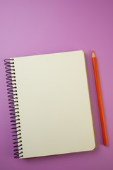 spiral-bound notebook and orange pencil lie on a lilac background.