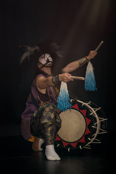 Taiko drummer in a wig and a demon mask on stage squarts with drumstick in his arms outstretched and taiko drum on a black background. Demon from Japanese mythology.