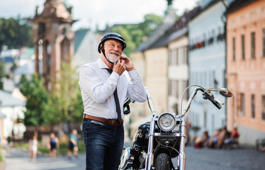 A senior businessman with motorbike in town, putting on helmet.