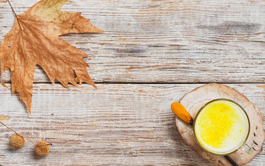 Cup with golden milk with turmeric on a wooden substrate. Next to a spoon with turmeric. Leaf and fruits of a plane tree against wooden background. Top view. Copy space