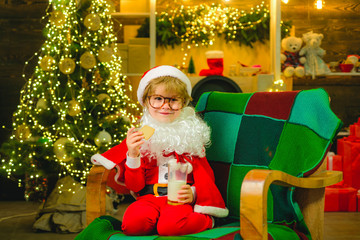 Fototapeta na wymiar Happy Santa Claus with glass of milk and cookie. Cheerful Santa Claus child holding glass with milk and cookie with fireplace and Christmas Tree in the background.
