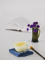Cup with golden milk on a blue napkin. Near a wooden spoon with turmeric. In the background is an open book and a bouquet of wildflowers in a vase. White background. Close-up.