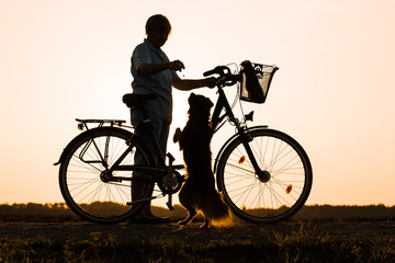 Fototapeta na wymiar Senior woman standing near a bike and feeding a dog, silhouette of person at sunset with pet, rest at a trip 
