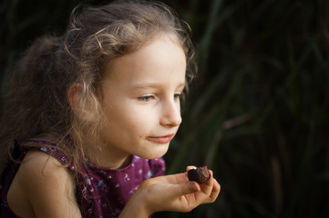 Beautiful little blond girl in violet dress with ponytail is eating a chocolate candy in the park durring a day, sugar addiction concept