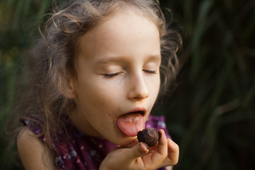 Beautiful little blond girl in violet dress with ponytail is eating a chocolate candy in the park durring a day, sugar addiction concept