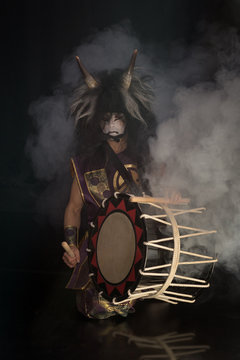 Taiko drummer in a wig and a demon mask on stage squarts with drumstick and drum on a black background with smoke. Demon from Japanese mythology.