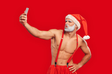 Christmas Freestyle. Young bearded Santa Claus bare muscular upper body in hat standing isolated on red taking selfie on phone pouting lips to camera silly