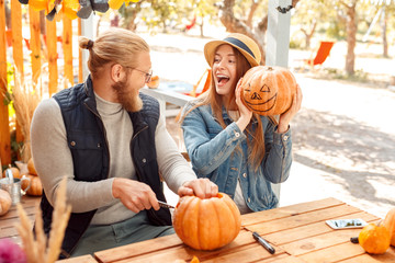 Halloween Preparaton Concept. Young couple sitting at table outdoors making jack-o'-lantern girl...