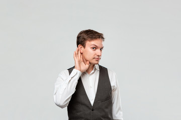 Handsome young man in a white shirt and grey vest holding hand close to ear listening to some interesting rumor.