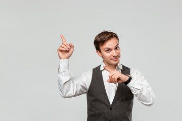 Handsome man in a white shirt and grey vest showing with his fingers at the empty space for advertising in the upper left corner of light grey background.