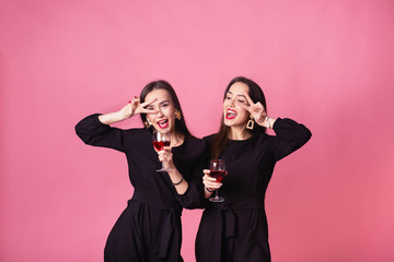 Two women celebrate the New Year party having fun laughing and drinking wine.   Girls dance and posing on pink background, cheerful emotions