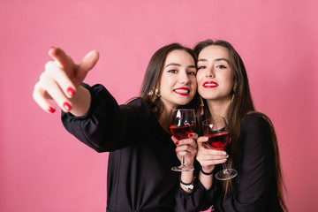 Two women celebrate the New Year party having fun laughing and drinking wine.  Girls posing and smiling on pink background, cheerful emotions, bright makeup, red lips, indoors party