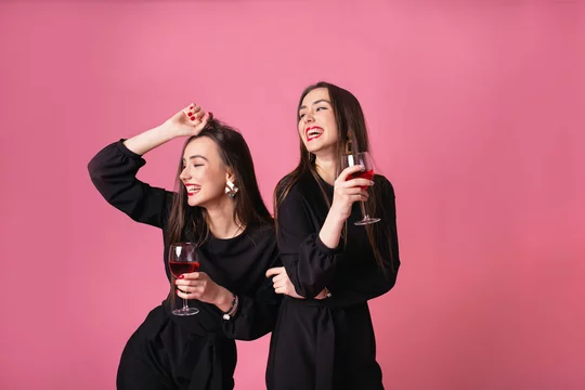 Two attractive women in night dress drinking wine isolated on a plastic  pink background. Girls posing and smiling, cheerful emotions, indoors  party, wearing black stylish evening outfits. Stock Photo | Adobe Stock