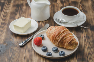 Delicious breakfast with fresh berries. Coffee cup, creamer and croissant with berries in white bowl and butter on wooden background.