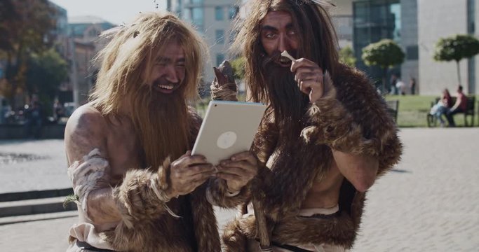 Close up neanderthals in animal fur using modern tablet browsing internet having fun city center caveman civilization computer device evolution future homo sapiens human hunter-gatherers slow motion