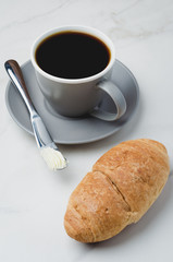 Coffee break with croissant, butter knife and cup of black coffee on a white stone table.