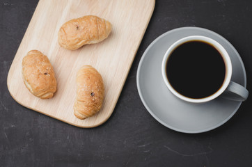 Grey cup with black coffee and wooden tray with croissants on dark table. Top view. Coffee break.