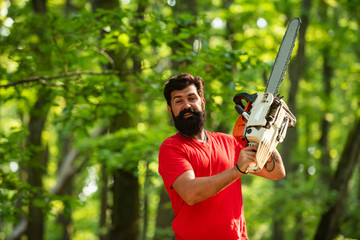 Agriculture and forestry theme. Lumberjack with chainsaw on forest background. Stylish young man posing like lumberjack.