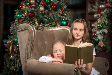 Boy and girl read a book while sitting in an armchair in front of a Christmas tree