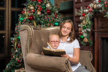 Boy and girl read a book while sitting in an armchair in front of a Christmas tree