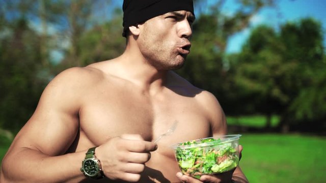 Young muscular sportsman eating healthy food outdoors and looking on camera. Fresh salad and carrot are rich in vitamins needed for developing strong muscular tissue. Naked sexy athlete preparing for