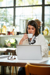 Attractive caucasian elegant brunette sitting in cafe, drinking coffee and looking at laptop. On table is laptop.
