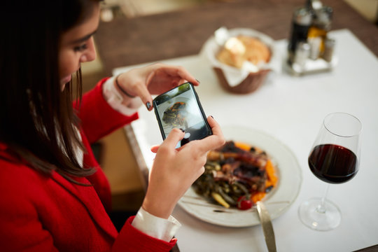 Top view of attractive businesswoman in suit sitting in restaurant and taking picture of food over smart phone.