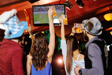 group or company of young people friends in Bavarian caps - guys and girls watching football on TV in a sports bar holding glasses with beer. Celebration of the Oktoberfest festival