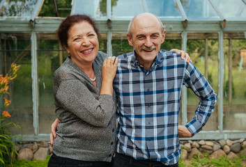 Smiling lovely old happy couple in front a glass greenhouse near summer house