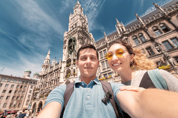 happy multinational couple in love hugs and takes a selfie photo on the background of The city hall tower in Munich. Honeymoon trip to Germany - obrazy, fototapety, plakaty