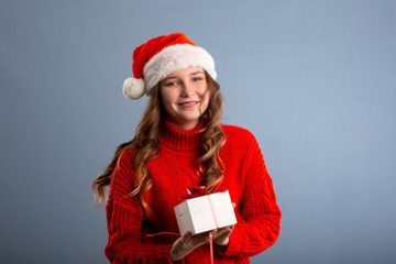 Beauty Christmas fashion model girl holding Xmas gift box. Beautiful lady, Long straight flying hair, red santa hat, holding gift box. Sexy young woman portrait isolated on gray background