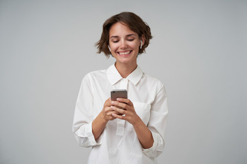 Cheerful young brunette lady with short haircut holding mobile phone in raised hands and looking happily at screen, wearing formal clothes while standing over white background