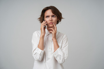 Serious looking young brunette woman with short haircut holding mobile phone and having conversation with her partners, frowning eyebrows musingly, isolated over white background