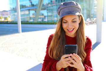 Happy beautiful girl using smart phone in modern city. Fashion autumn winter girl portrait.