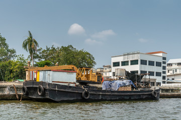 Fototapeta na wymiar Bangkok city, Thailand - March 17, 2019: Bangkok Noi Canal. Small freighter barge with yellow crane and housing on top docked along the canal under blue sky. White building in back. 