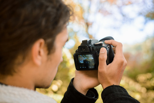 Portrait of male photographer shooting video in live-view mode of beautiful colorful autumn forest, holding camera in front of his face, eager to catch perfect moment