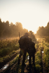Rifle Hunter and His Son Silhouetted in Beautiful Sunset. Huntsman with a boy and rifle in a forest on a sunrise. - obrazy, fototapety, plakaty