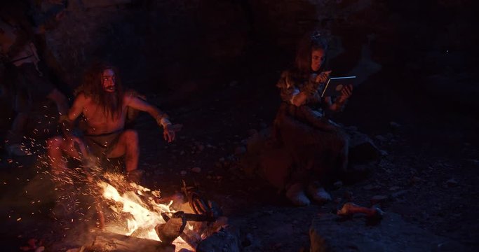 Slow motion tribe cavemen neanderthals wearing animal skin use tablet computer sitting by the fire in a cave stone fun male ancient history evolution nature homo sapiens primitive campfire