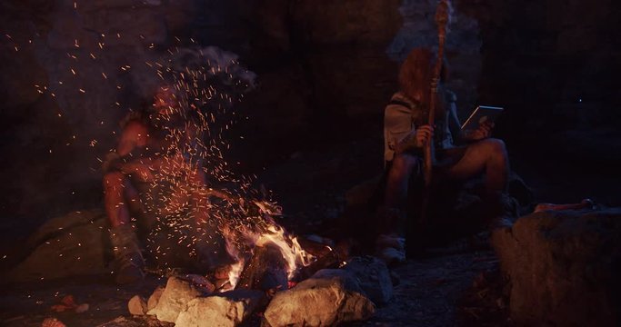 Slow motion tribe cavemen neanderthals wearing animal skin use tablet computer sitting by the fire in a cave stone fun male ancient history evolution nature homo sapiens primitive campfire clos eup