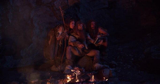 Close up cavemen hold bone with meat neanderthals wearing animal skin use tablet computer waving his hand having fun sitting by the fire in cave stone ancient evolution homo sapiens slow motion