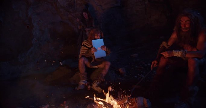 Close up cavemen neanderthals wearing animal skin discovering tablet computer sitting by the fire in a cave stone fun ancient history portrait evolution homo sapiens primitive campfire slow motion