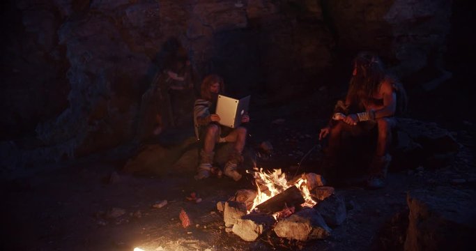 Close up neanderthals wearing animal skin discovering tablet computer sitting by the fire in a cave stone fun male ancient history portrait evolution nature homo sapiens primitive campfire slow motion