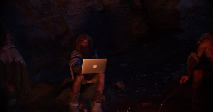 In dark neanderthals wearing animal skin use tablet computer sitting by the fire at night in a cave stone fun male ancient history evolution nature homo sapiens primitive campfire slow motion
