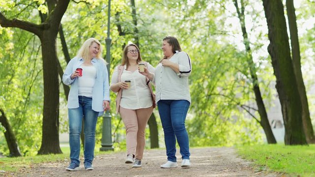 Full length shot of group of three middle aged plus size women walking down path in green park with takeaway coffee cups, talking joyfulle and laughing